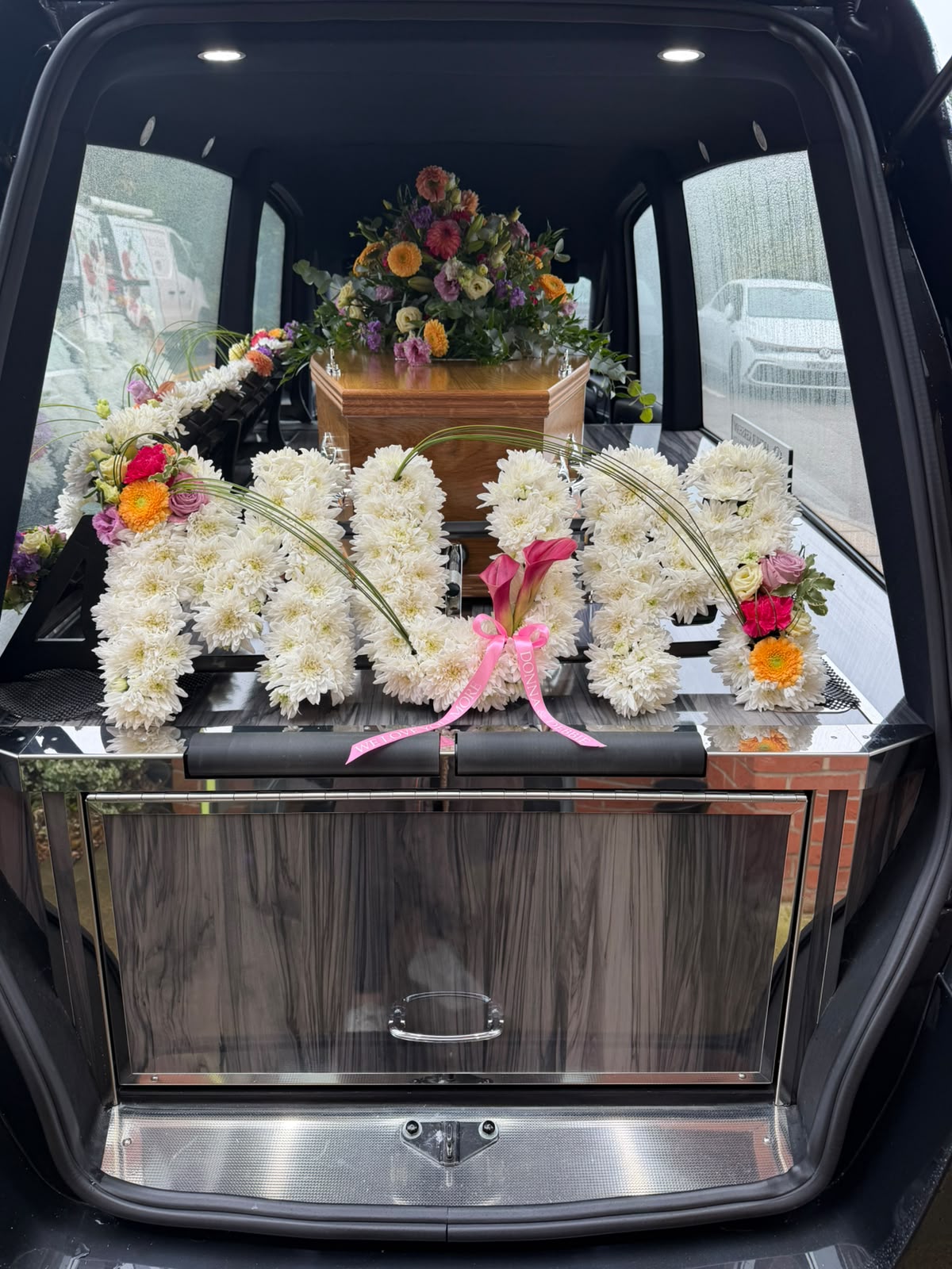 Personalised white floral letter tribute with colourful floral accents displayed beside a coffin