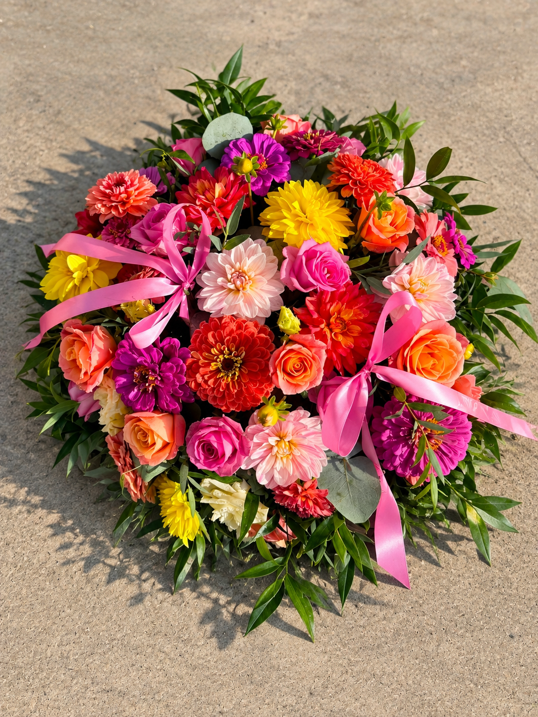 Round funeral floral tribute in vibrant pink, orange and yellow flowers with pink ribbons